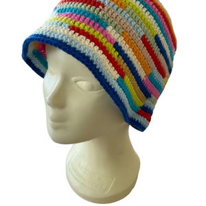 Op de afbeelding: Een veelkleurige gehaakte vissershoed. De hoed heeft een levendige mix van regenboogkleuren, waaronder rood, geel, blauw en roze, in een gestreept patroon. De rand heeft een blauwe rand. De hoed wordt getoond op een mannequinhoofd.