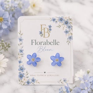 Puede incluir: Un par de pendientes de botón con forma de pequeñas flores de color azul claro de cinco pétalos y centro amarillo. Los pendientes están sujetos a una tarjeta expositora blanca con el texto Florabelle Bloom y Real Flower Jewellery impreso.
