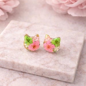 Puede incluir: Un par de pendientes de botón con forma de gato hechos de resina transparente. Cada pendiente contiene flores secas incrustadas en tonos rosa vibrante y verde brillante, acentuadas con pequeñas motas de pan de oro en el material.