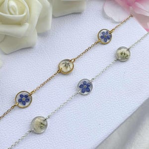 May include: Two bracelets, one gold and one silver, each with clear, circular pendants. The pendants contain pressed flowers, some with blue blossoms and others with white. The bracelets are displayed on a white surface, with floral accents.