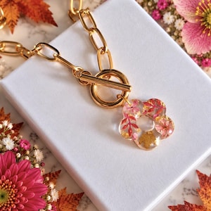 May include: A gold-toned chain necklace featuring a toggle clasp and a flower-shaped pendant made of clear resin. The pendant contains embedded pink and red dried flower petals along with small gold-coloured flakes throughout the material.