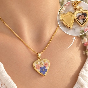 Pode incluir: Um colar com relicário em forma de coração dourado com fundo branco, flores prensadas cor-de-rosa e azuis e uma letra S dourada. O relicário abre-se para revelar um compartimento interior circular com uma fotografia de um casal.