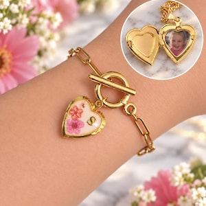 May include: A gold-toned chain bracelet featuring a heart-shaped charm with pressed pink and orange flowers and the letter S. An inset shows an open gold heart-shaped locket containing a photograph of a child, attached to a gold chain.
