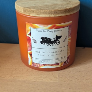 Peut inclure: Une bougie cylindrique orange dans un pot en verre avec un couvercle en bois clair. L'étiquette présente un graphique de char noir, le texte Vivi's Tea Party présente : HELIOS LE BRILLANT Bougie artisanale et des tranches d'oranges sanguines.