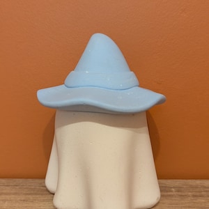 Peut inclure: Un chapeau de sorcière bleu clair coiffe une figurine blanche en forme de fantôme. Le chapeau a un large bord et un sommet pointu. Le fantôme a une base arrondie et une forme drapée, créant une décoration sur le thème d'Halloween.