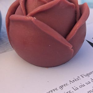 May include: A dusty rose-colored candle shaped like a blooming rose with a single white wick centered at the top. The wax petals are sculpted in a layered, rounded form, resting on a white surface with printed French text visible at the bottom.