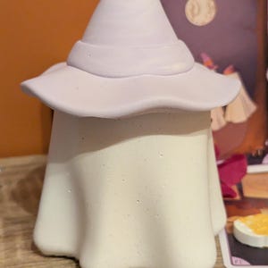 Peut inclure: Figurine de fantôme en céramique avec un chapeau de sorcière. Le fantôme est blanc avec une surface texturée, et le chapeau est violet clair. Le chapeau a un large bord et un sommet pointu. Décoration sur le thème d'Halloween ou de la fantaisie.