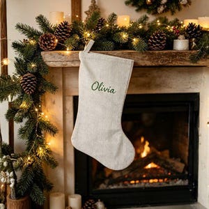 Peut inclure: Une chaussette de Noël en lin beige clair avec le prénom Olivia brodé en écriture cursive vert foncé. La chaussette possède une boucle simple sur le dessus pour la suspendre et présente un design épuré en tissu tissé texturé.