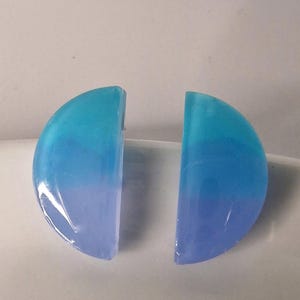 Puede incluir: Dos pendientes en forma de semicírculo con un degradado de tonos azules. Los pendientes transitan de un lavanda claro a un turquesa vibrante. Los pendientes probablemente están hechos de resina o un material similar.