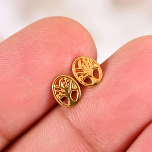 May include: A pair of small, gold-coloured stud earrings. Each earring has a tree design within an oval shape. The earrings are shown against a light background.