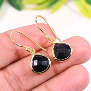 May include: A pair of gold-coloured drop earrings featuring black, faceted, teardrop-shaped gemstones. The earrings have a simple, elegant design with a hook closure. The gemstones are set in a gold-coloured bezel setting.