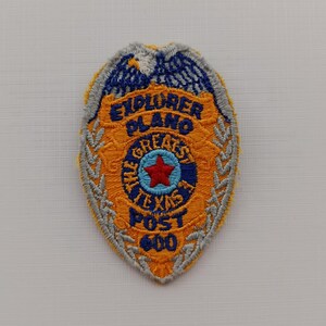 Può includere: Toppa ricamata a forma di distintivo. La toppa è principalmente arancione con dettagli blu e argento. Il testo "EXPLORER PLANO GREATEST TEXAS POST 600" è ricamato sulla toppa, insieme a una stella rossa.