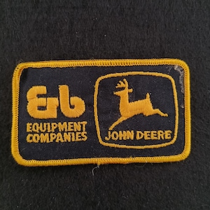 Peut inclure: Un écusson rectangulaire avec un fond bleu marine et une bordure dorée. Le côté gauche présente les lettres "Eb" en or, avec "EQUIPMENT COMPANIES" en dessous. Le côté droit affiche un cerf doré et les mots "JOHN DEERE".
