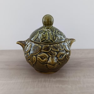 Op de afbeelding: Een groene keramische pot met deksel en een gestructureerd, bladvormig patroon. De pot heeft een afgeronde vorm met kleine handvatten en een knop op het deksel. De kleur is een gedempt olijfgroen.