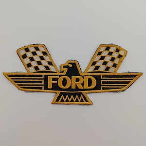 Pode incluir: Patch bordado com um design de águia estilizada em preto e dourado, com a palavra "FORD". As asas da águia são bandeiras quadriculadas em preto e branco. O patch tem um fundo preto e acabamento dourado.