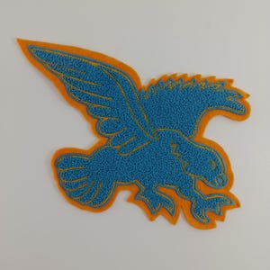 Könnte beinhalten: Gestickter Aufnäher in Form eines fliegenden Adlers. Der Adler ist blau mit goldenen Details und einem orangefarbenen Rand. Der Aufnäher ist wahrscheinlich für eine Jacke oder eine Tasche.