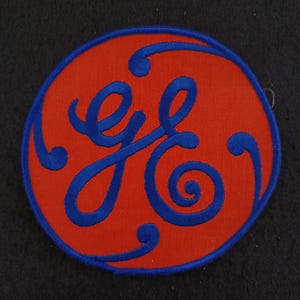 Pode incluir: Um patch redondo vermelho com uma borda azul e um logotipo "GE" azul estilizado no centro. O logotipo apresenta linhas fluidas e cursivas e redemoinhos decorativos. O patch é provavelmente um emblema ou uma insígnia.