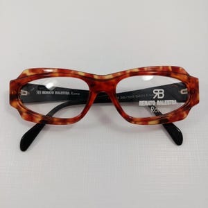 Pode incluir: Óculos com armação retangular com padrão tartaruga. Os óculos têm hastes pretas e lentes transparentes. A marca "RENATO BALESTRA Roma" está impressa na armação. O tamanho da armação é 39/525 54017-140.