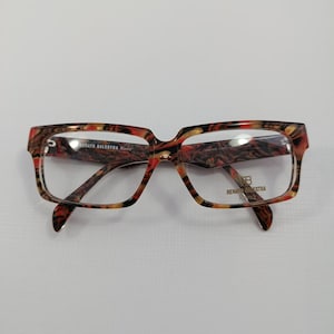 Pode incluir: Óculos retangulares com padrão tartaruga em tons de castanho, laranja e preto. As armações têm um design espesso e arrojado com lentes transparentes. A marca "Renato Balestra Roma" é visível.
