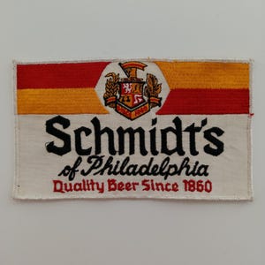 Pode incluir: Patch bordado com o logotipo da Schmidt's of Philadelphia. O patch tem um esquema de cores vermelho, amarelo e branco com as palavras "Quality Beer Since 1860" em vermelho.