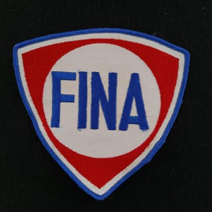 Könnte beinhalten: Ein Vintage-Patch in Schildform, mit einem weißen Kreis mit den blauen Buchstaben "FINA". Der Kreis ist von einem roten Rand umgeben, und der gesamte Patch ist blau umrandet. Der Patch befindet sich auf einem schwarzen Hintergrund.