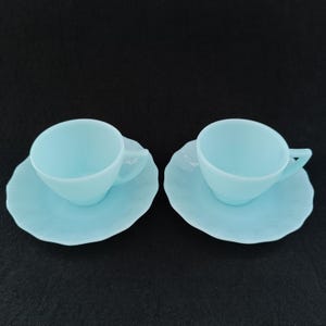 Op de afbeelding: Twee bijpassende lichtblauwe kopjes en schotels. De kopjes hebben een eenvoudig oor en staan op een geschulpte schotel. De set is gemaakt van een solide, ondoorzichtig materiaal. De achtergrond is effen zwart.