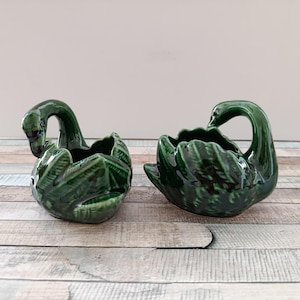 Puede incluir: Dos maceteros con forma de cisne de cerámica en un tono verde intenso y brillante. Los maceteros tienen texturas de plumas detalladas y cuellos curvos, creando un diseño decorativo. Se colocan sobre una superficie de madera.