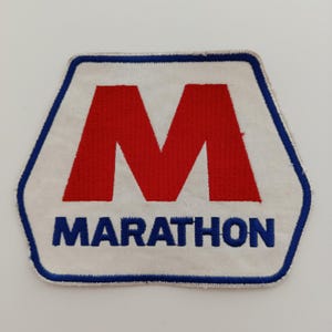 Pode incluir: Um patch hexagonal branco com uma borda azul. O patch apresenta um grande "M" vermelho no centro e a palavra "MARATHON" em azul na parte inferior. O patch é provavelmente um emblema ou logotipo.