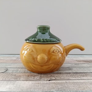 Pode incluir: Jarro de cerâmica amarelo com um design de rosto sorridente e tampa verde. Possui uma alça lateral. A tampa tem o texto "EAR SCO". O jarro está sobre uma superfície de madeira.