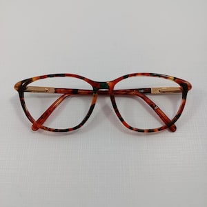 Può includere: Occhiali di forma ovale con motivo tartarugato nei toni del marrone, arancione e nero. Le montature hanno dettagli metallici color oro sulle aste e sul ponte. Le lenti sono trasparenti.