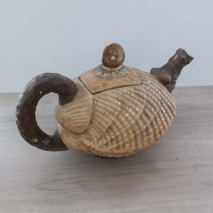 Könnte beinhalten: Keramik-Teekanne mit einem strukturierten, muschelartigen Design in Beige- und Brauntönen. Der Griff und der Ausguss sind dunkelbraun, und der Deckel hat einen kleinen, runden Knopf. Die Teekanne steht auf einer hellen Holzoberfläche.