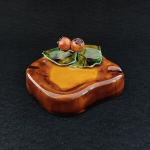 Pode incluir: Um cinzeiro de cerâmica com acabamento castanho brilhante. O cinzeiro tem formato quadrado com bordas arredondadas e uma depressão central. Duas pequenas frutas redondas marrom-alaranjadas estão sobre folhas verdes. É um item decorativo.