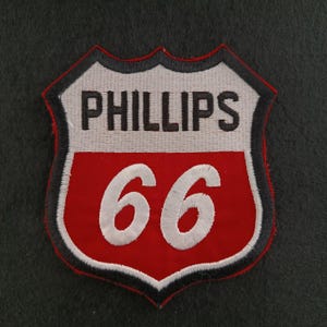 Op de afbeelding: Geborduurde patch met het Phillips 66 logo. De patch heeft de vorm van een schild met een wit bovenstuk en een rood onderstuk. De woorden "PHILLIPS" en "66" zijn prominent aanwezig.