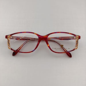 Pode incluir: Um par de óculos com armação vermelha e estampada. As lentes retangulares são transparentes e as hastes têm um detalhe dourado. A marca "NEOSTYLE" está impressa na armação.