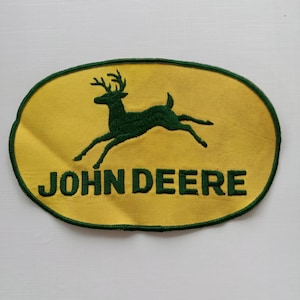 Op de afbeelding: Geborduurde patch met het John Deere-logo. De patch heeft een gele ovale achtergrond met een groene rand. Een groen springend hert staat in het midden, met de groene woorden "JOHN DEERE" eronder.