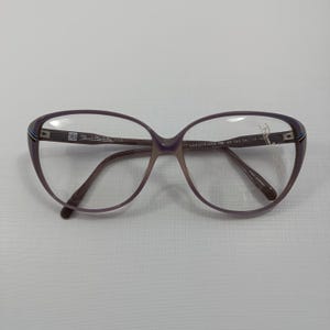 Pode incluir: Um par de óculos vintage com armação roxa e lentes transparentes. Os óculos têm formato de olho de gato e hastes marrons. O texto "GREGORIANA RB 43 744 58 14-14" está impresso na armação. Fabricado na Alemanha.