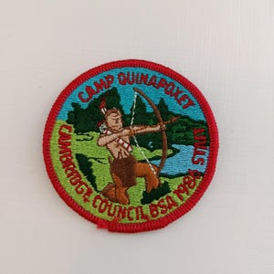 Pode incluir: Um patch bordado redondo com uma borda vermelha. O patch apresenta uma figura nativa americana com arco e flecha, em um cenário verde. O texto "CAMP QUINAPOXET" e "CAMBRIDGE COUNCIL BSA 1986 STAFF" estão em um padrão circular.