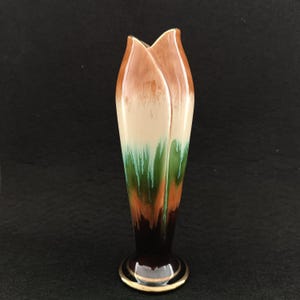 Puede incluir: Un jarrón alto con forma de tulipán y parte superior acampanada. El jarrón presenta un degradado de colores, comenzando con marrón en la parte superior, pasando a crema, verde, naranja y terminando con una base marrón oscuro con borde dorado.