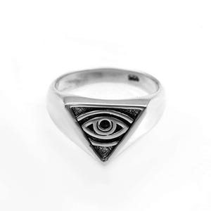 Op de afbeelding: Een zilverkleurige ring met een driehoekig voorvlak voorzien van een gegraveerd oogsymbool. Het oogontwerp is gecentreerd in de driehoek en omringd door donkere, verdiepte details die contrasteren met het gepolijste, gladde oppervlak van de ring.