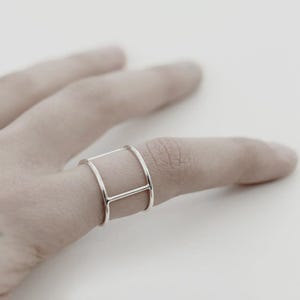 May include: A polished silver ring featuring a wide, open-frame design. The band consists of two parallel circular hoops connected by a single vertical bar on one side, creating a structured, geometric appearance that encircles the finger.