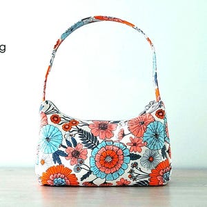 May include: A white baguette bag with a floral print featuring orange, blue, and pink flowers. The bag has a single strap and is made of fabric.