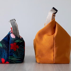 May include: Two fabric bags, one with a tropical print and the other a solid orange color. Both bags have a single handle and are folded into a square shape.
