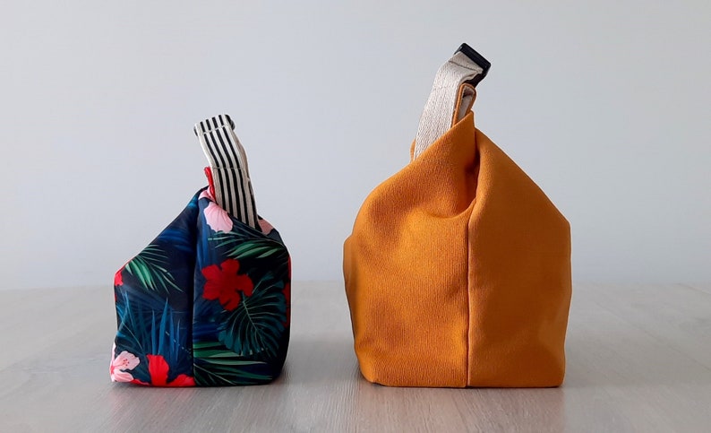 May include: Two fabric bags, one with a tropical print and the other a solid orange color. Both bags have a single handle and are folded into a square shape.
