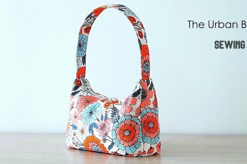 May include: A white fabric handbag with a floral print in shades of orange, pink, and blue. The bag has a single top handle.
