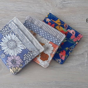 May include: Four different fabric card wallet sewing patterns with floral designs. The patterns are folded and stacked on a light wood surface. The text "EKO CARD WALLET SEWING PATTERN Story Quilt Etsy Shop" is printed on the surface.