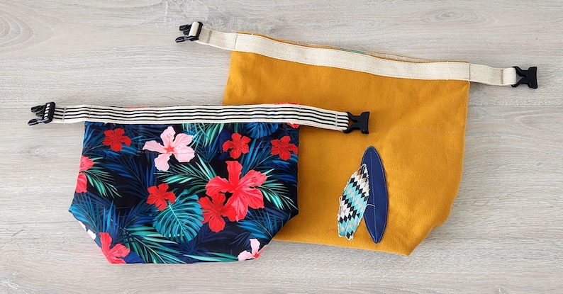 May include: Two reusable shopping bags. One bag is yellow with a blue and white surfboard design. The other bag is black with a tropical floral print featuring pink and red flowers.