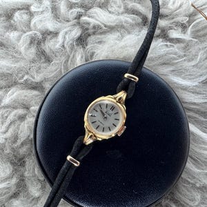 Op de afbeelding: Een vintage goudkleurig polshorloge met een lichtgekleurde wijzerplaat en zwarte wijzers. Het horloge heeft een zwarte leren band en wordt gepresenteerd op een zwart oppervlak. Een label met een wapen is zichtbaar op de achtergrond.