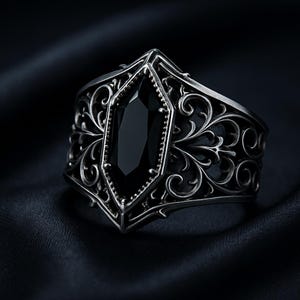 May include: A silver-toned ring with an ornate design. The ring features a large, black, rectangular gemstone at its center, surrounded by intricate scrollwork. The band is wide and detailed, creating a bold statement piece.