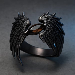 Könnte beinhalten: Ein dunkelgrauer Ring mit detaillierten Flügeldesigns auf beiden Seiten, die einen braunen, oval geformten Stein einrahmen. Der Ring hat ein strukturiertes, metallisches Aussehen, das auf ein gotisches oder Fantasy-Thema hindeutet.
