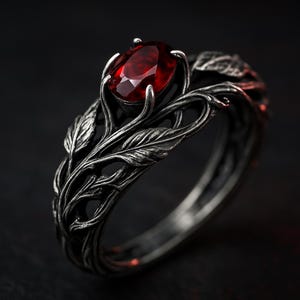 May include: A silver ring with a deep red, oval gemstone. The ring has a detailed, nature-inspired design with leaves and vines. The gemstone is held by four prongs.